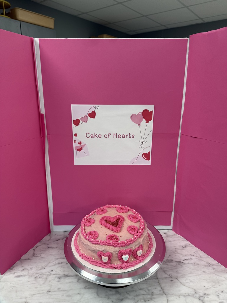 Pink frosted “Cake of Hearts” decorated with heart-shaped designs and a large center heart, set in a red and pink display.