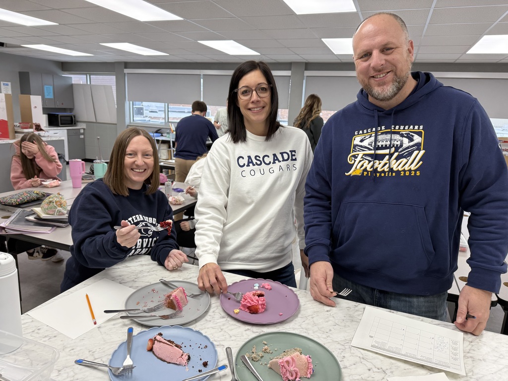 Three smiling staff members in Cascade Cougars gear taste Valentine-themed cakes in a classroom setting.