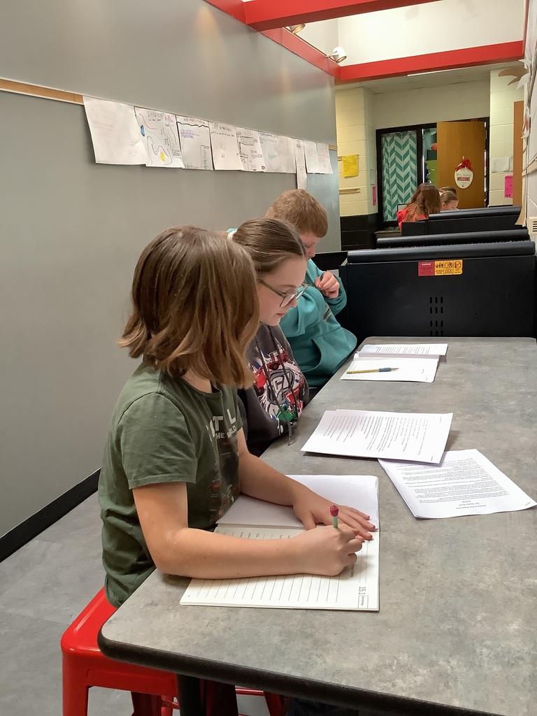Three students seated at a high table in a hallway, writing and discussing papers.