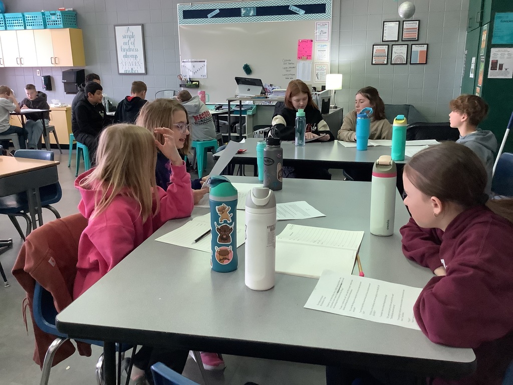 Students gathered around classroom tables, working in groups on writing assignments.