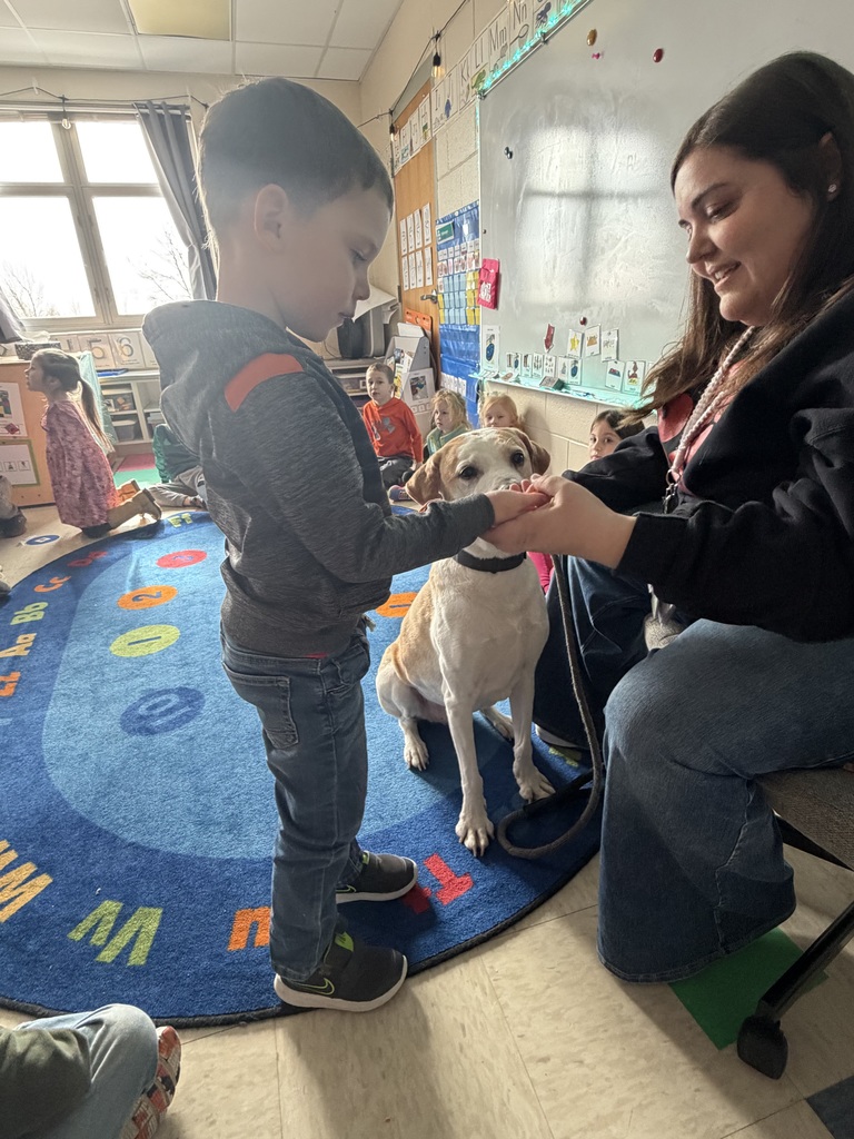 Student feeds a sitting dog while a teacher holds the leash and smiles.