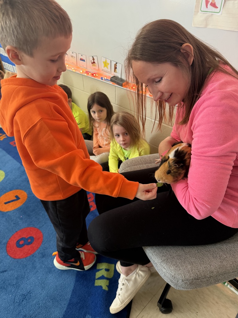 Teacher holds a guinea pig while a student feeds it, surrounded by other students sitting on a rug.