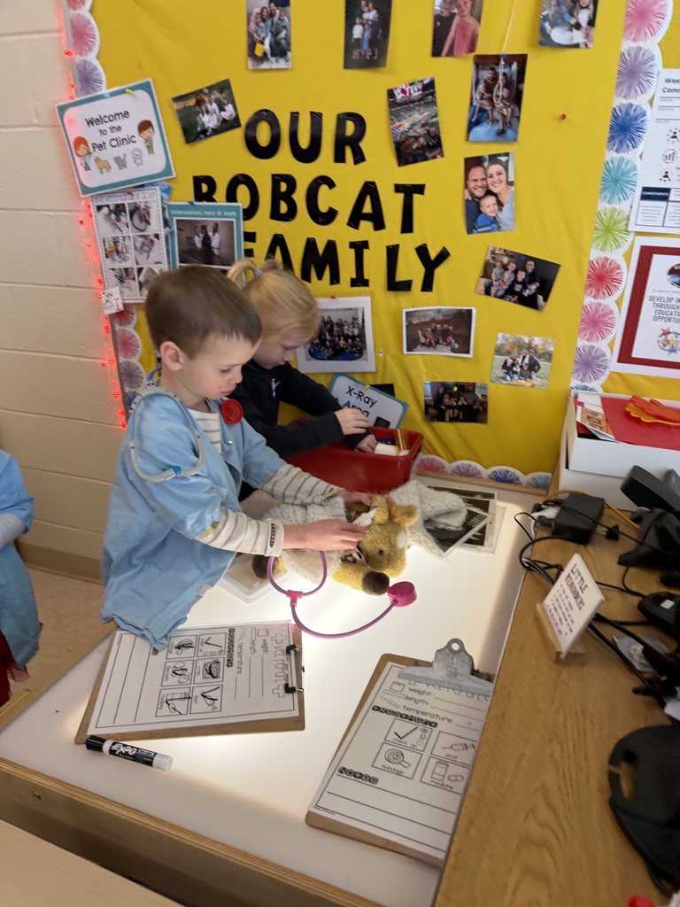 Two children in blue coats play pretend at a classroom pet clinic with a stuffed animal and toy tools.