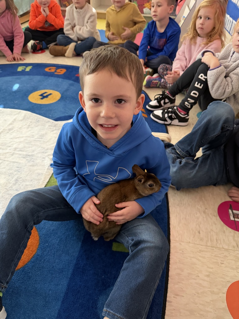 Young boy sits cross-legged on a classroom rug holding a brown rabbit as classmates watch.