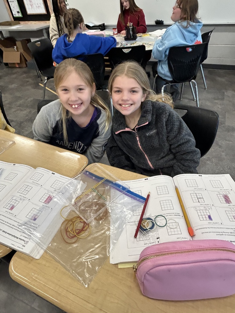 Two students smile at the camera while completing a math assignment with rubber bands and grid paper.