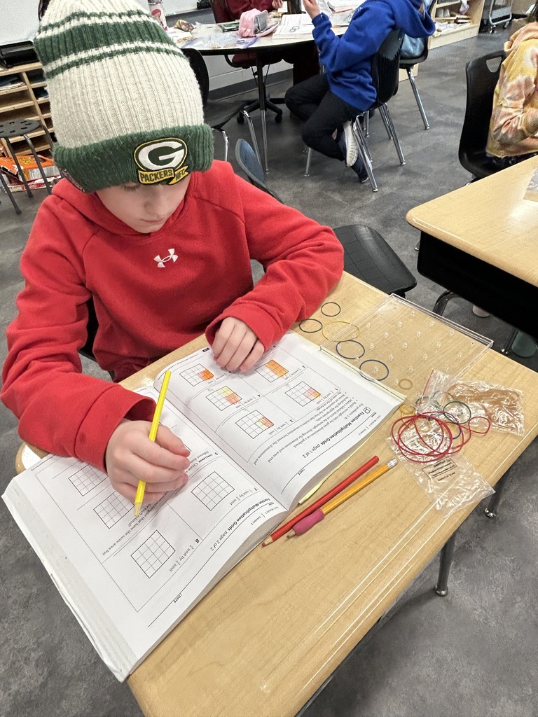 Student wearing a Packers hat colors grid boxes in a math workbook using pencils and rubber bands.