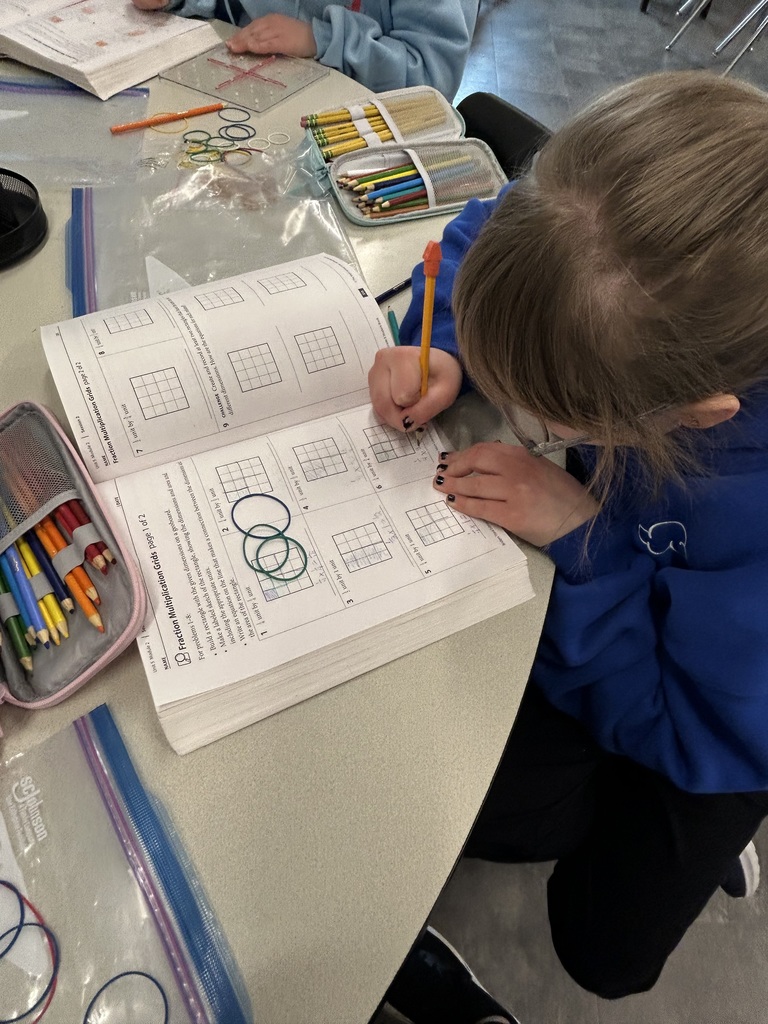 Student focuses on a math worksheet involving grids and colored rubber bands at a round table.