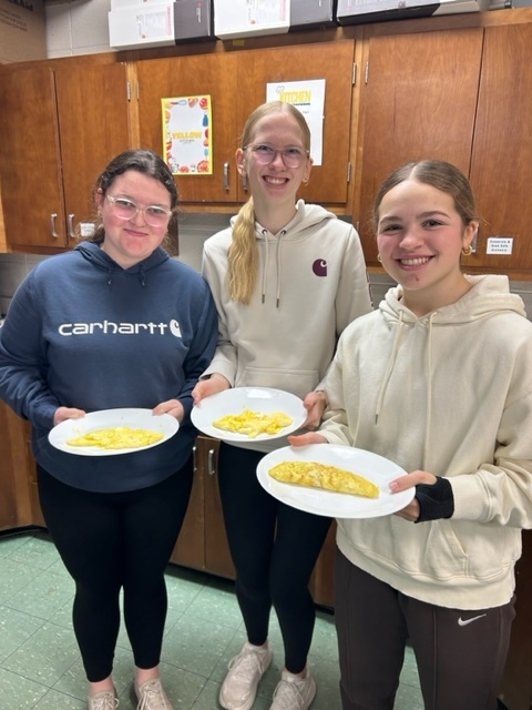 Three students smile while holding plates of cooked eggs in a kitchen classroom.