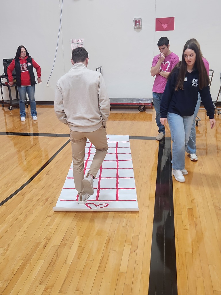 Students participate in an activity on a heart-decorated grid mat in a gymnasium.