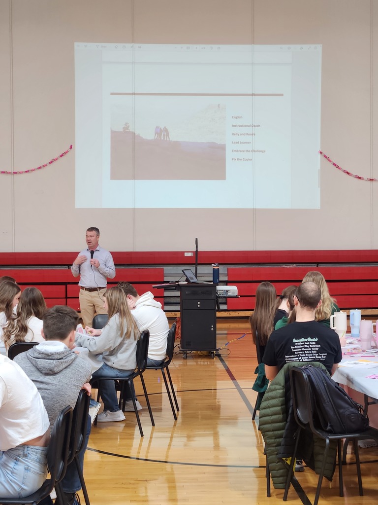 A man gives a presentation in a gym while students seated at tables listen and take notes.