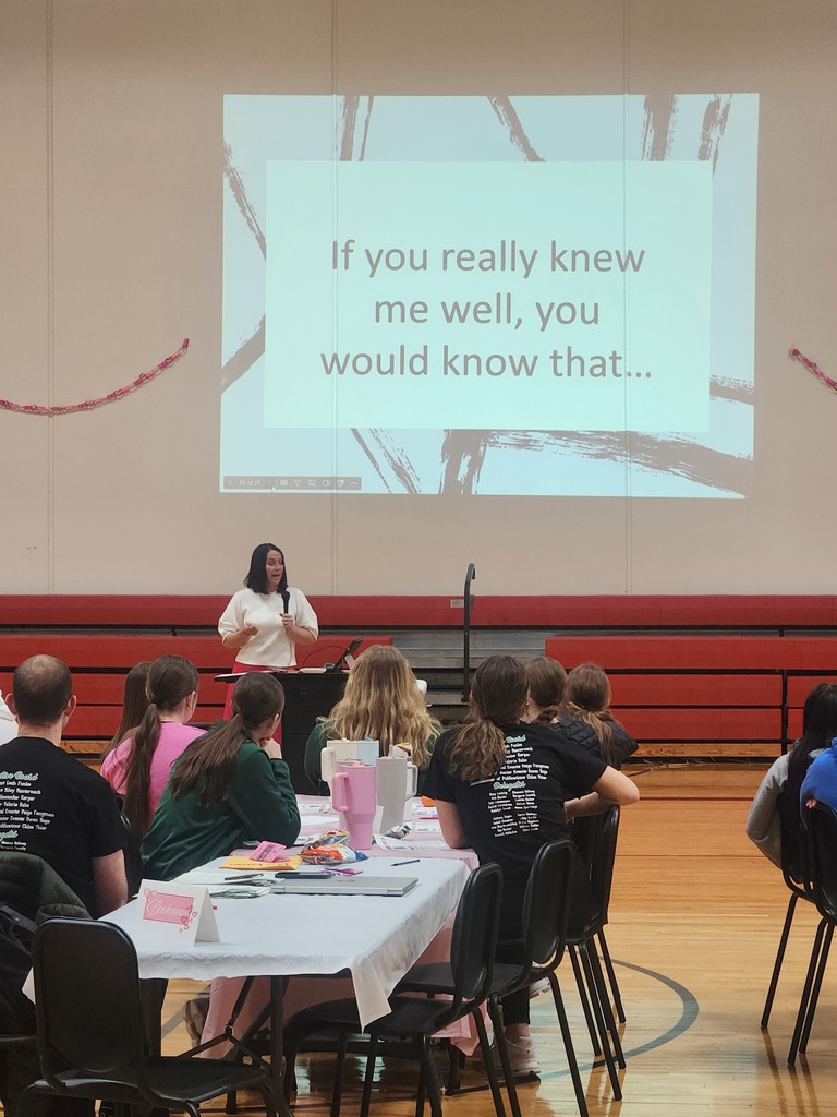A woman speaks to a group of students seated at tables, with a presentation slide that reads, “If you really knew me well, you would know that…”
