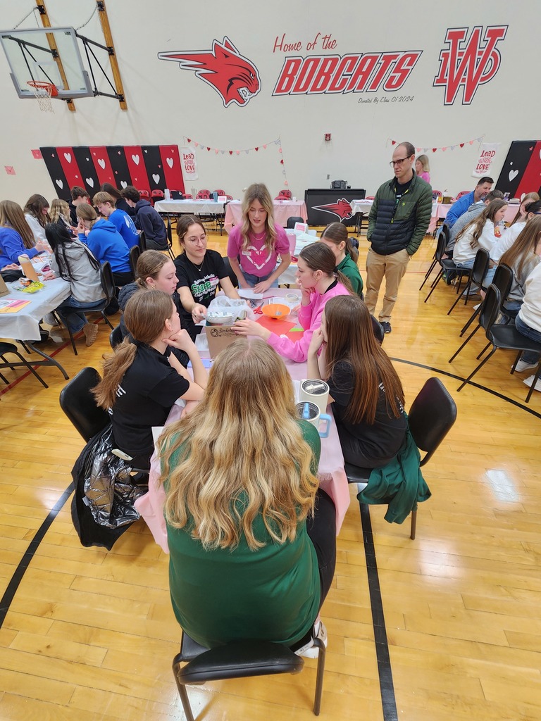 Students sit around a table working on an activity as others observe in a gym decorated with hearts and school logos.