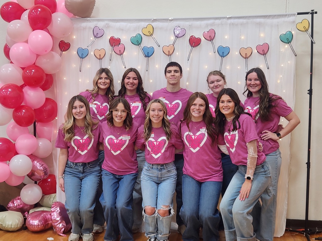 A group of students in matching pink heart shirts pose in front of a Valentine-themed backdrop with balloons and heart decorations.