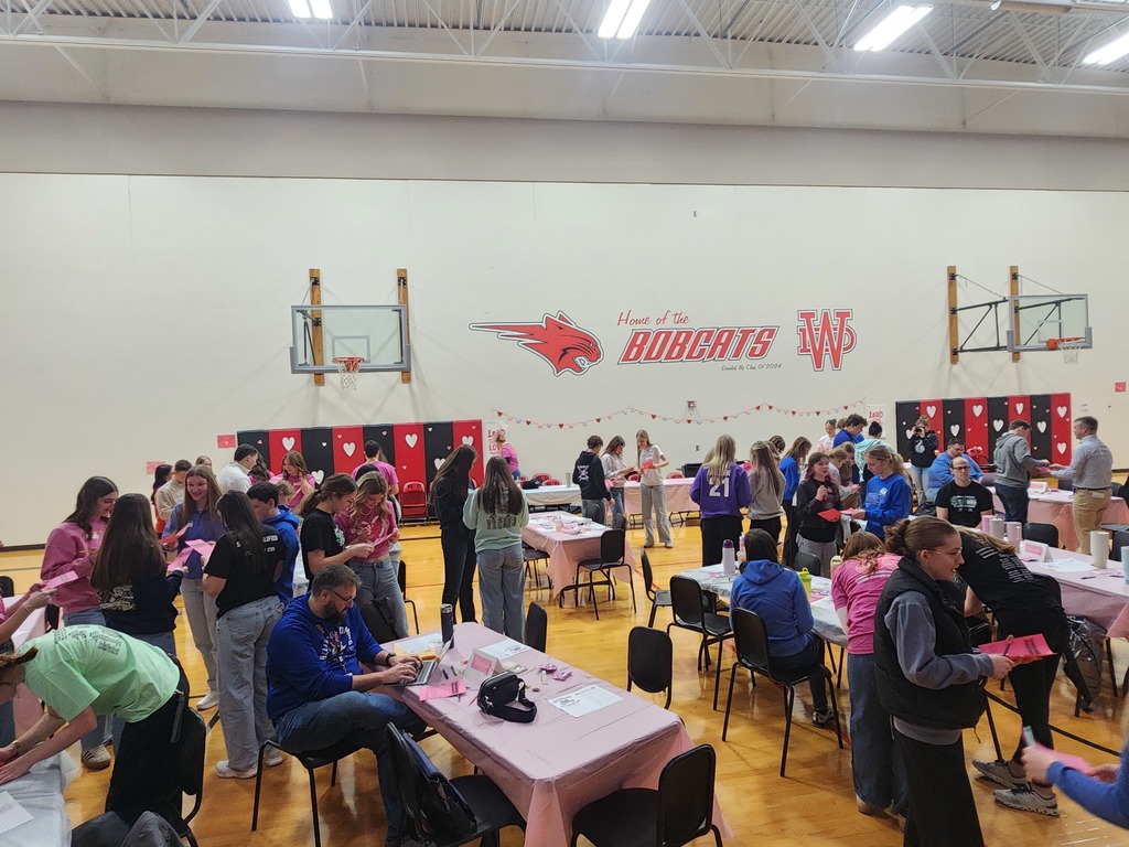 Large group of students gathers in a gym decorated for Valentine’s Day, working at tables with craft supplies.