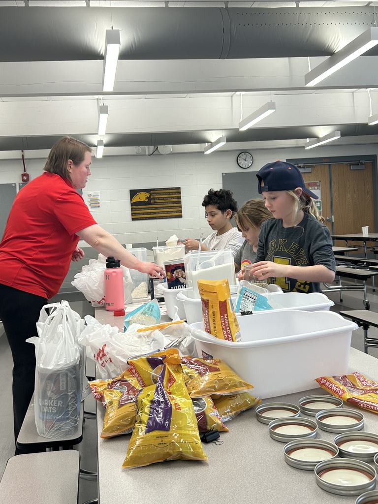 A group of students, guided by an adult, carefully measure and scoop ingredients into jars as part of a classroom cooking activity.
