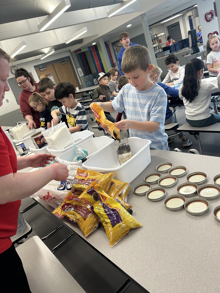 Students work together at a cafeteria table to fill jars with dry ingredients like oats and chocolate chips for a recipe project.