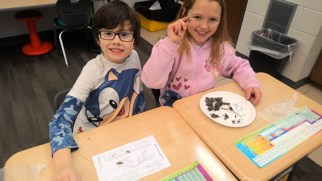 A student holds up a small item and smiles next to a classmate as they analyze materials on a plate.