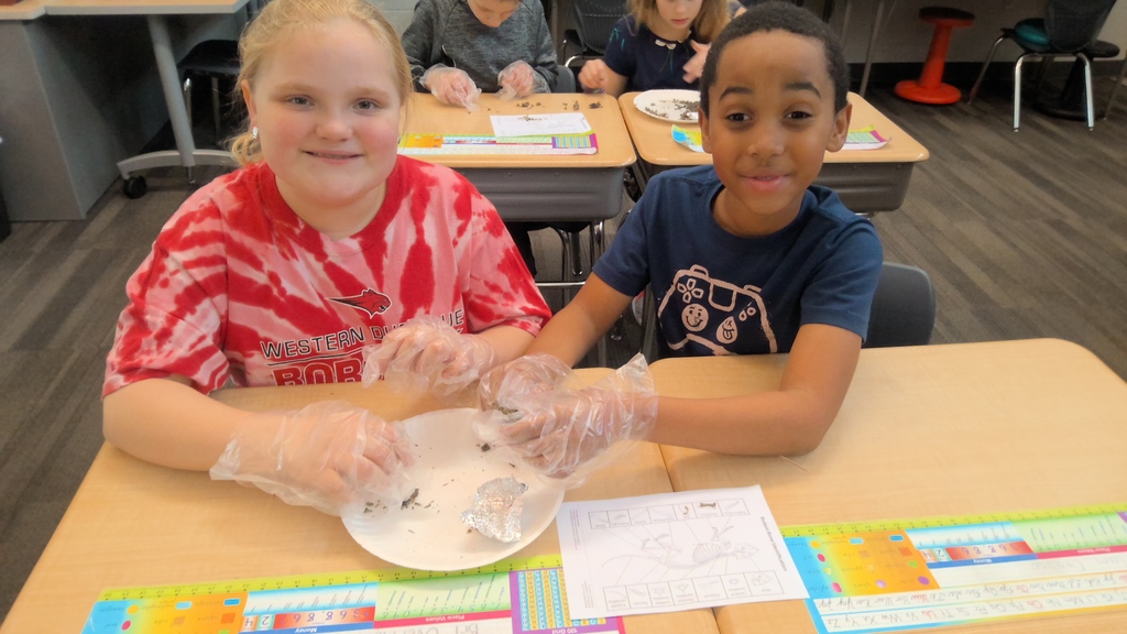 Smiling students work together at a desk, sorting through materials wearing plastic gloves.