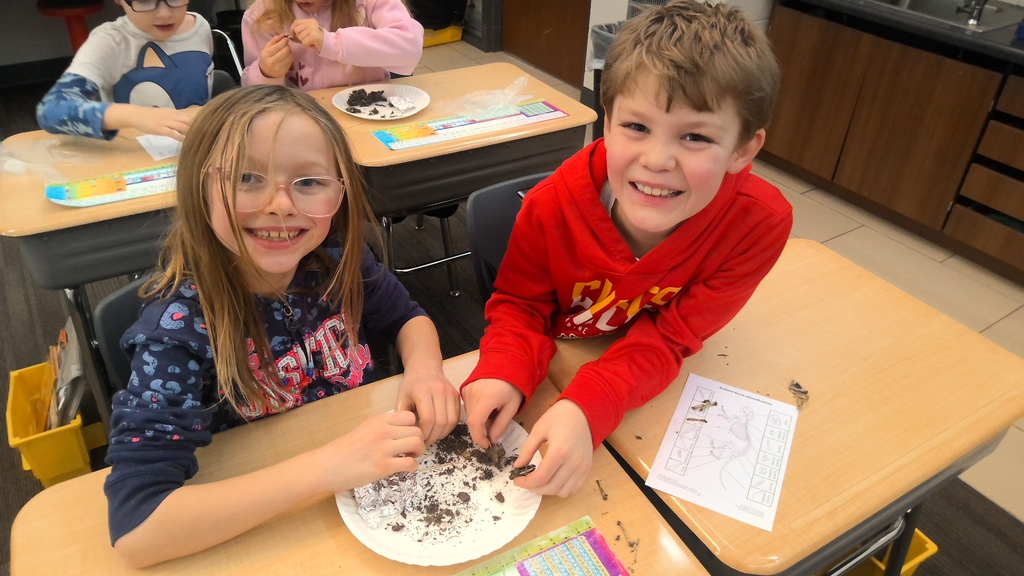 Two students work together and smile while sorting small objects on a plate during class.