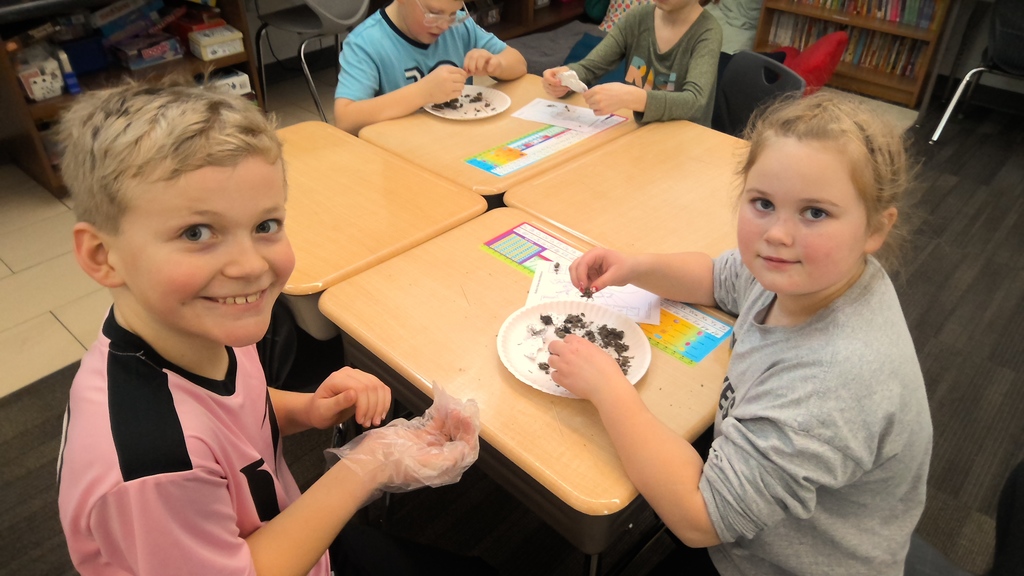 Two students smile at the camera while examining materials on a paper plate at their desks.