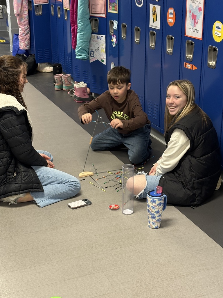 Two older students sit on the floor with a younger student building a structure with balancing sticks near school lockers.