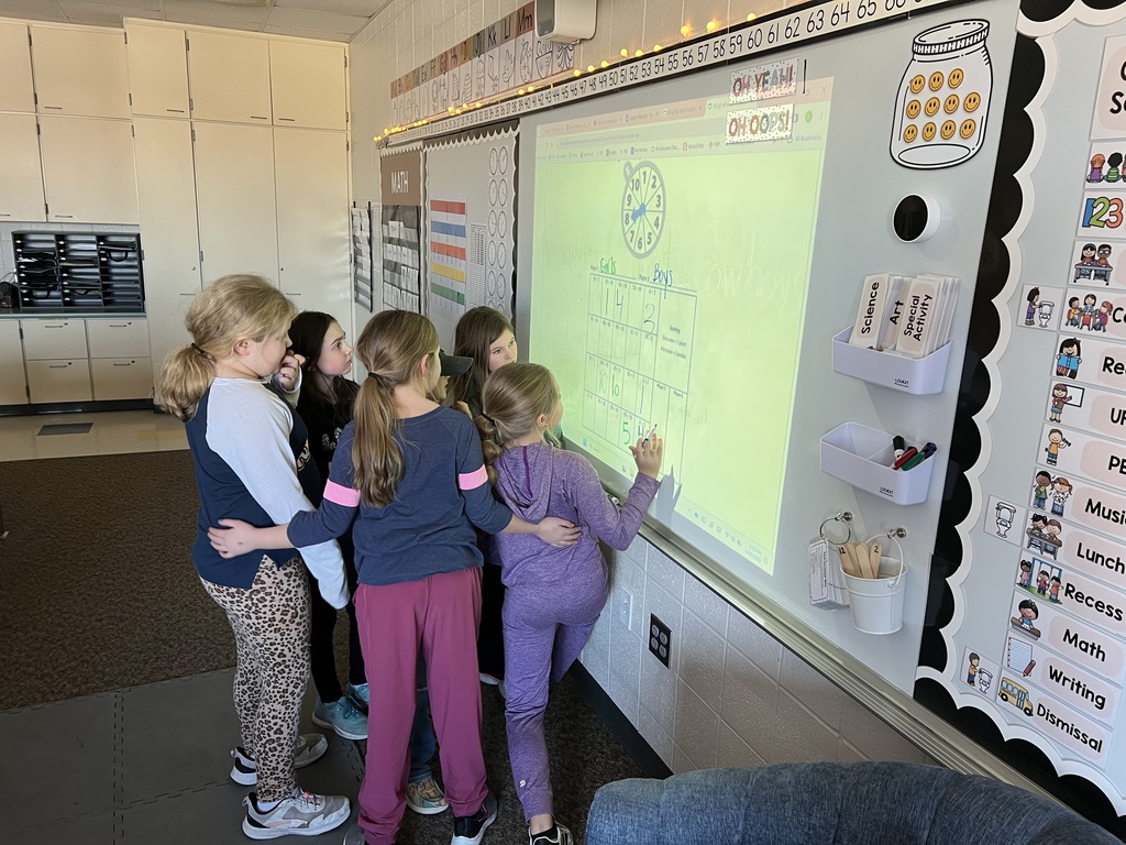 A group of young students gathers around an interactive whiteboard, collaborating on a classroom activity involving a chart labeled "Girls vs. Boys" with a clock at the top.