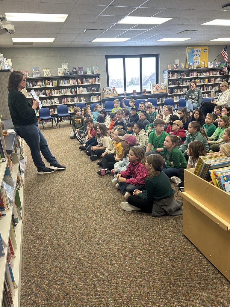A large group of young students sits attentively on the library floor while a woman at the front reads from a book, surrounded by bookshelves.