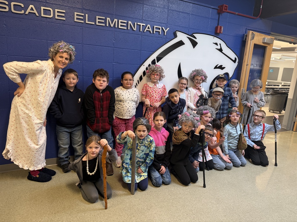 Group of children and one adult in costume pose in front of a Cascade Elementary wall display.