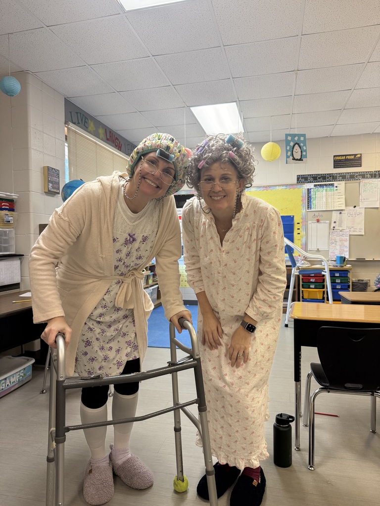 Two adults in costumes with robes, wigs, and glasses pose in a classroom, one using a walker.