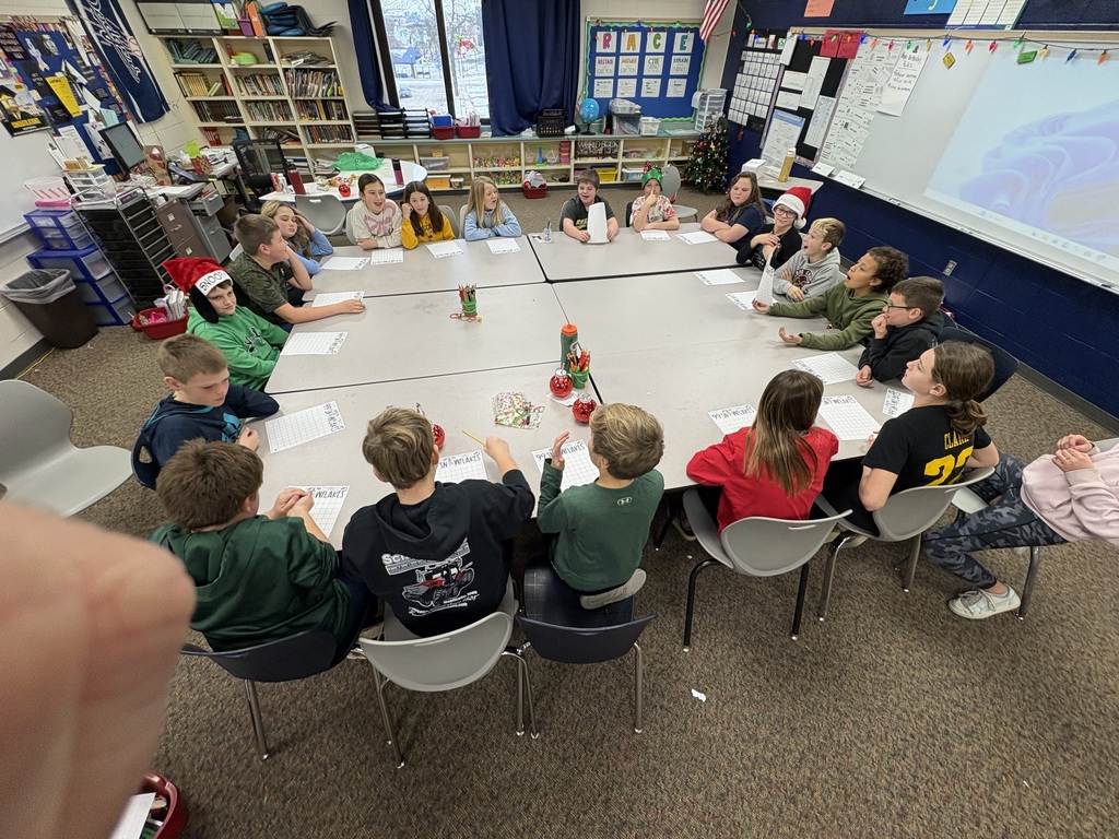 A class of students wearing festive clothing sit around a large group of tables, playing a holiday-themed game.