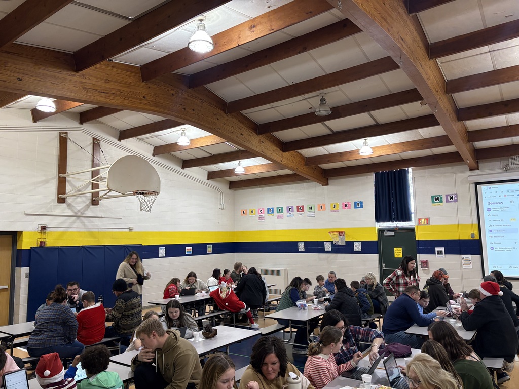 Families and students gather at cafeteria tables working on activities together in a school gymnasium decorated for the holidays.