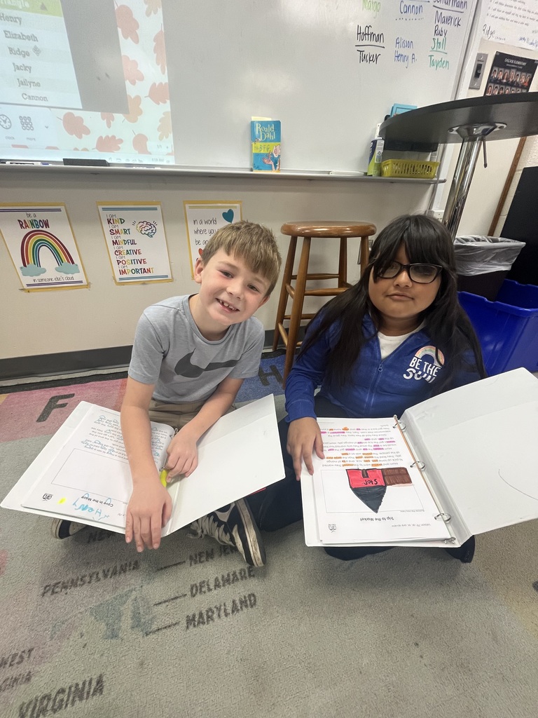 Two elementary students sit on the classroom floor smiling, each holding an open binder with colorful schoolwork displayed.