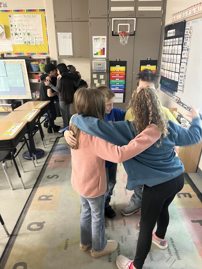 Groups of students in a classroom participate in a team-building activity, standing in small circles with arms around each other.