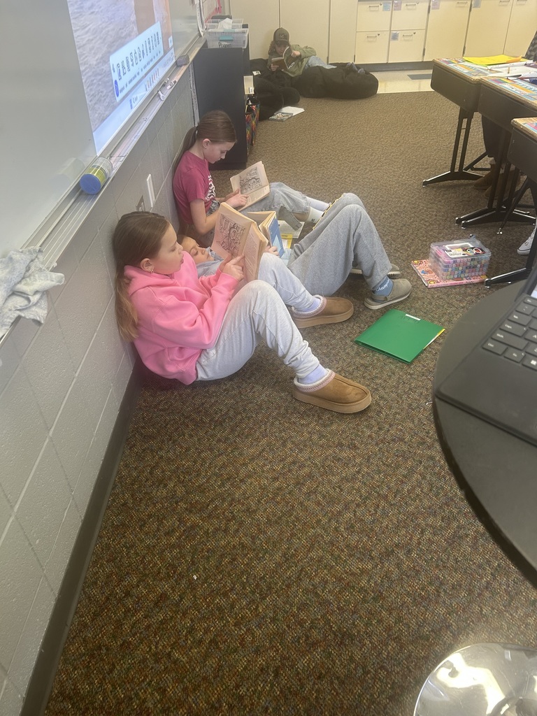 Students sit comfortably on the classroom floor, reading books independently. Some lean against the wall while others relax on bean bags in the back, focused on their reading.