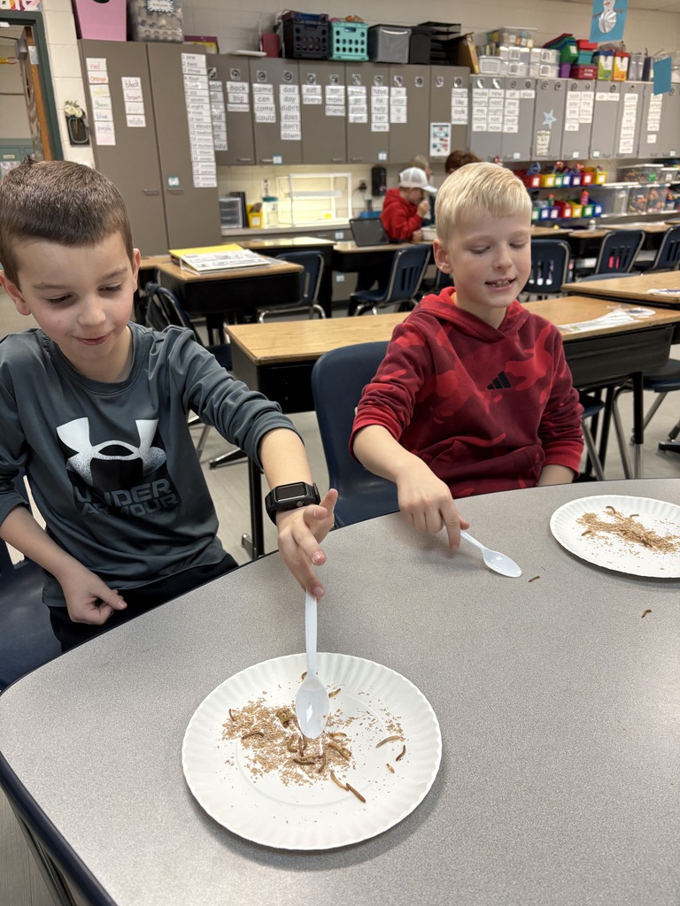Two students smile while using plastic spoons to explore worms on plates, participating in a hands-on science lesson.