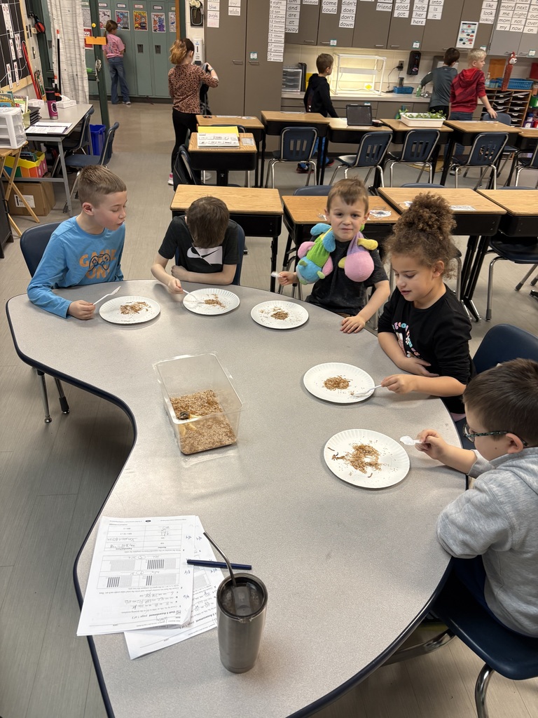 Students gather around a kidney-shaped table using plastic spoons to examine worms on paper plates during a classroom science activity.