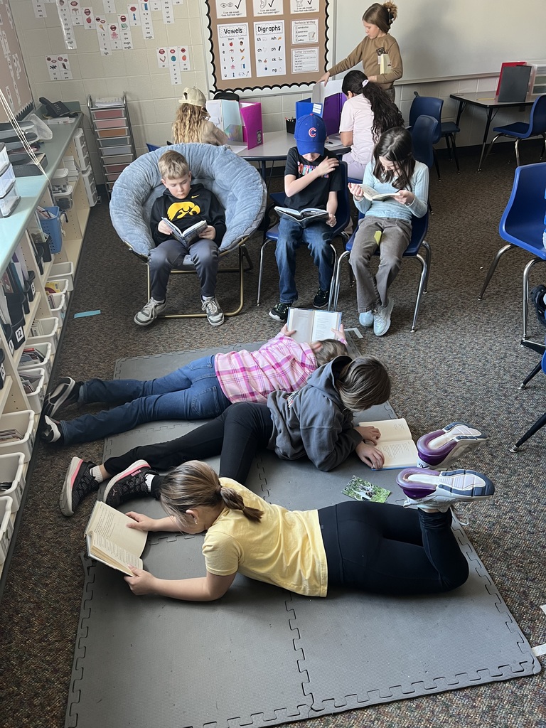 Students are spread out across the classroom reading books, with some lying on the floor and others seated in chairs.