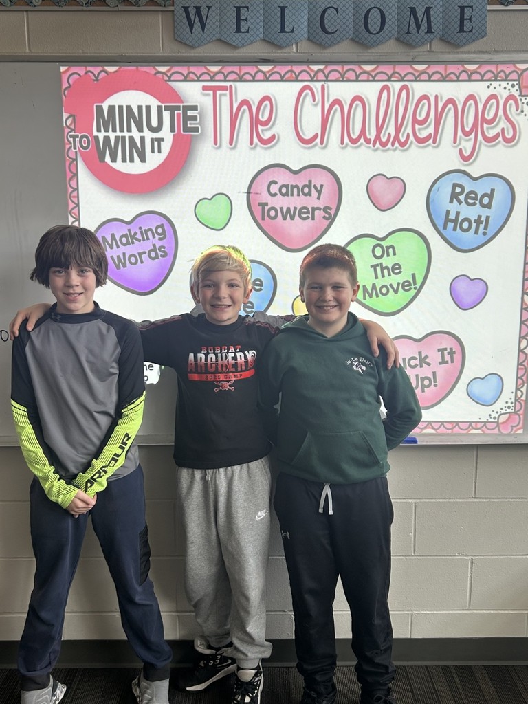 Three boys stand arm in arm smiling in front of a “Minute to Win It” classroom challenge board with colorful heart labels.