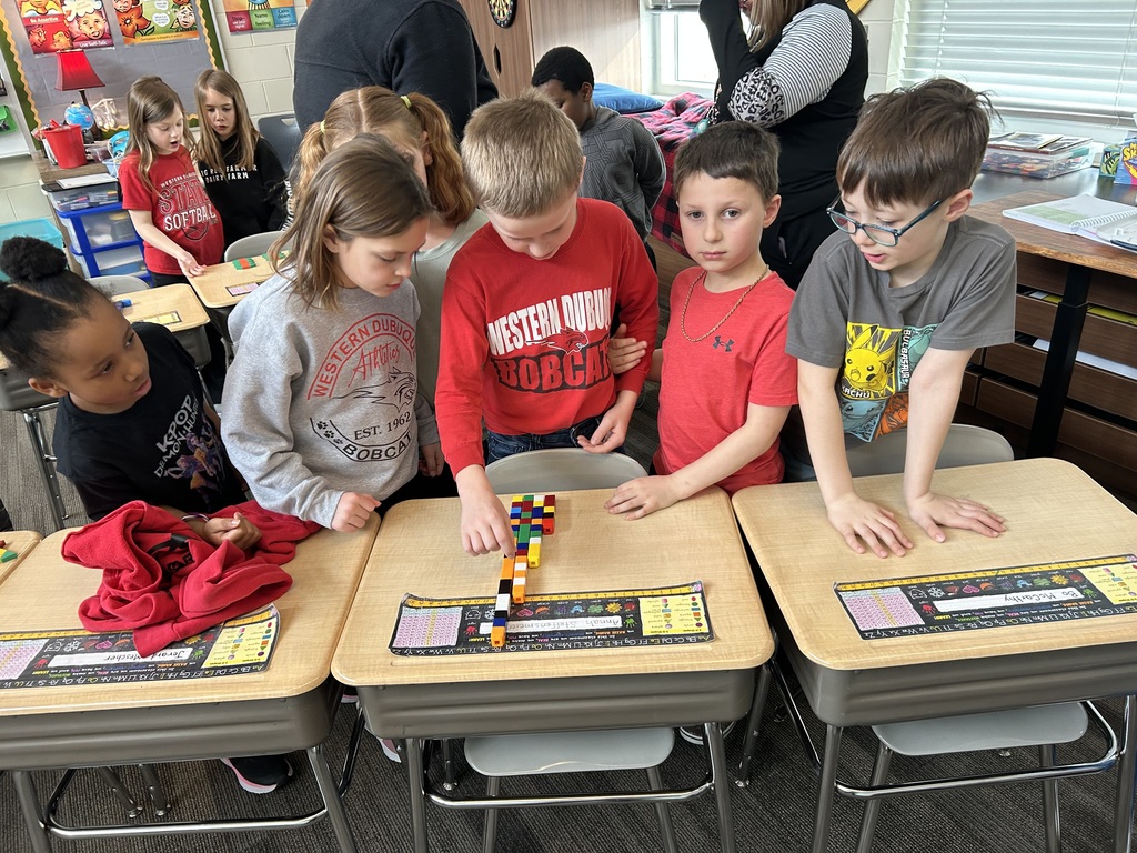Several students gather around desks, working together to build a tall structure with linking cubes.