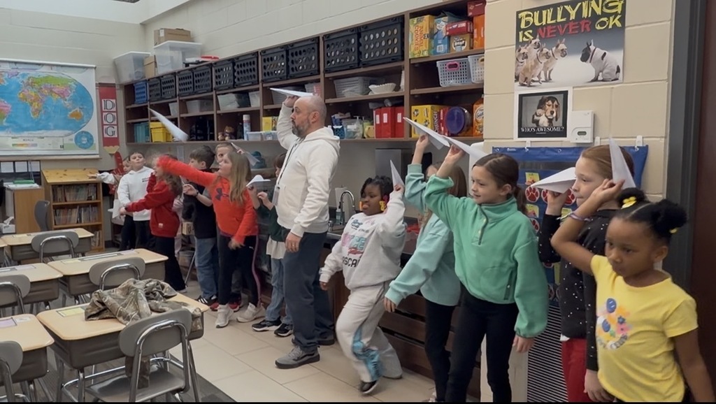 Students line up beside their teacher, holding paper airplanes and preparing to test their designs in class.