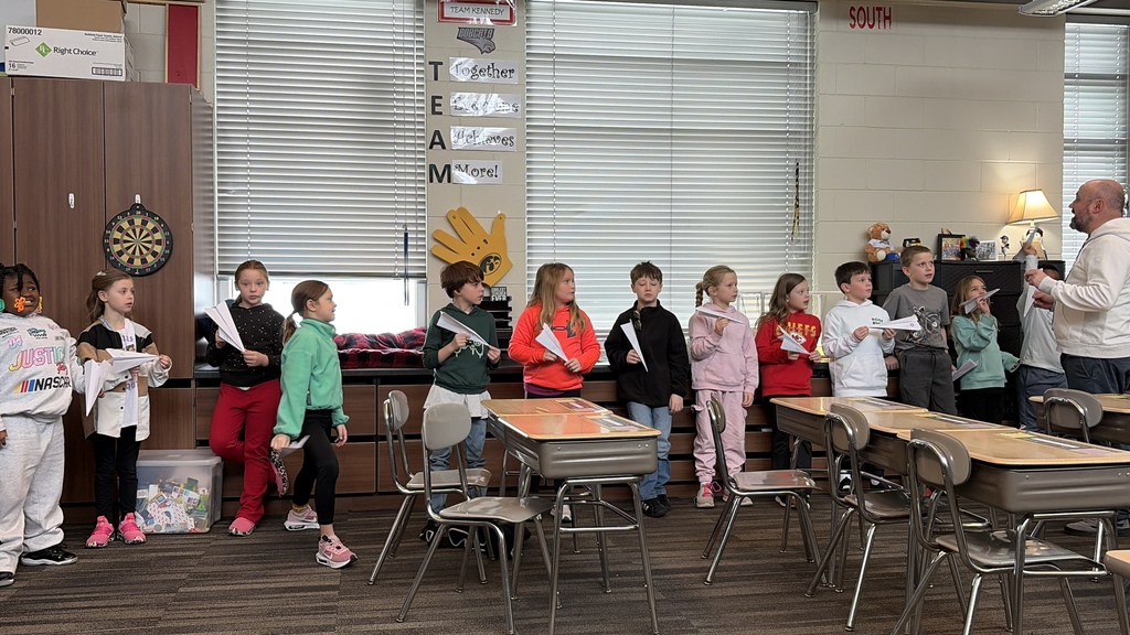 Teacher gives instructions as students stand in a row holding paper airplanes in a classroom activity.