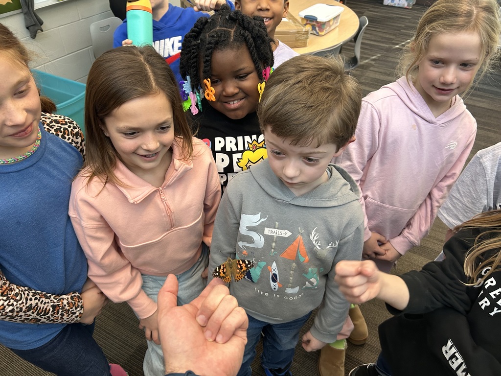 Group of students gather closely, smiling and watching a butterfly resting gently on a teacher’s hand in the classroom.