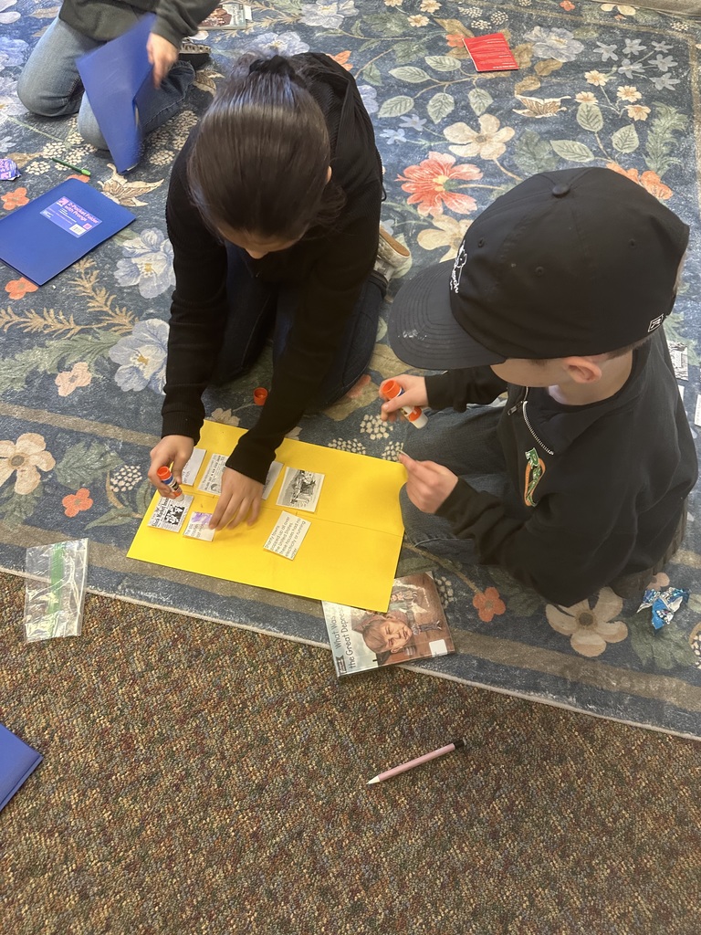 Two students sit on a classroom rug, gluing historical event cards onto a bright poster board for a timeline project.