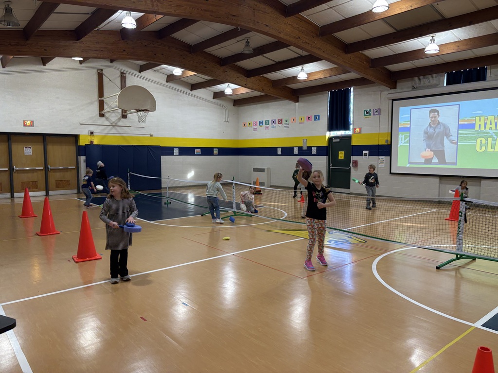 Students play paddle games over low nets in the gym while following instructions on a projected screen.