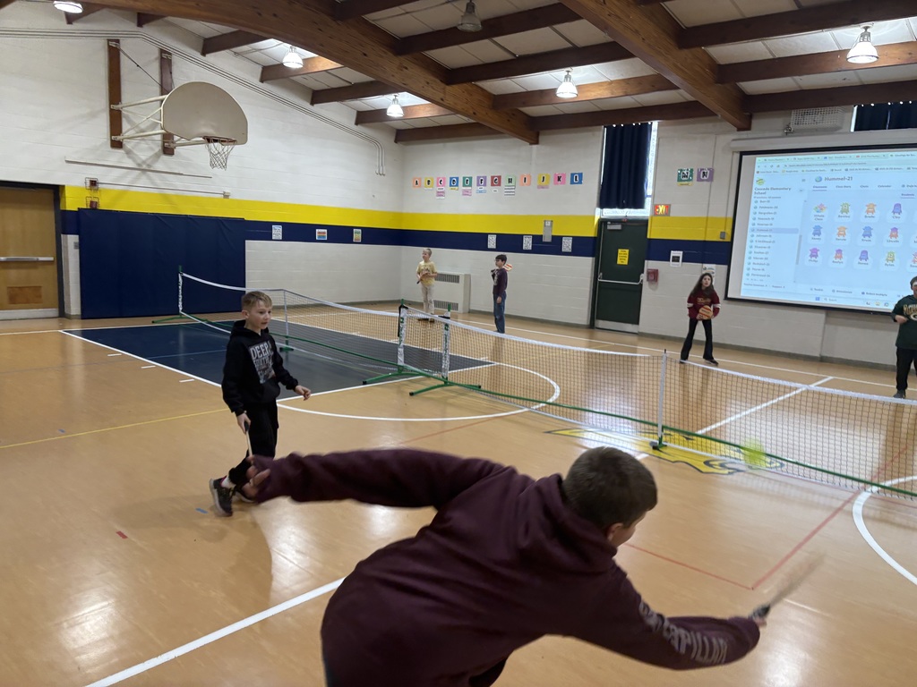 Student swings a paddle to hit a ball across the net as classmates wait their turn in the gym.
