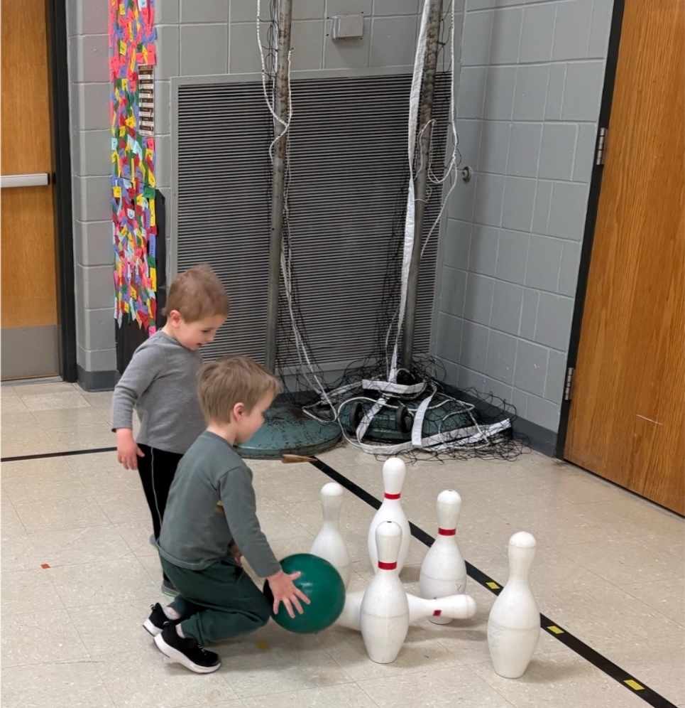 Two young students roll a green bowling ball toward large white pins set up in the hallway.