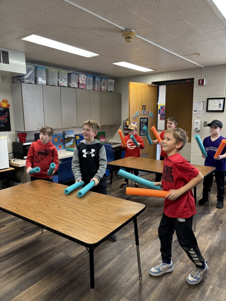 Students in a classroom smiling and moving with teal and orange pool noodles between tables during a group activity.