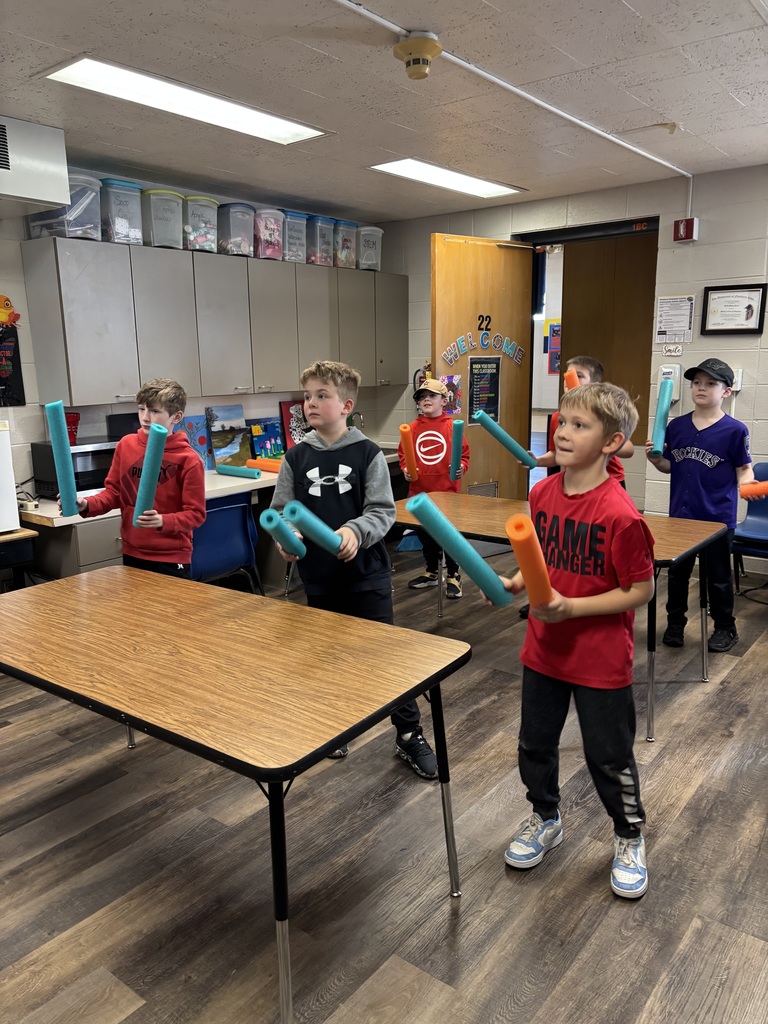 Elementary students standing between classroom tables holding colorful pool noodles during an interactive activity.