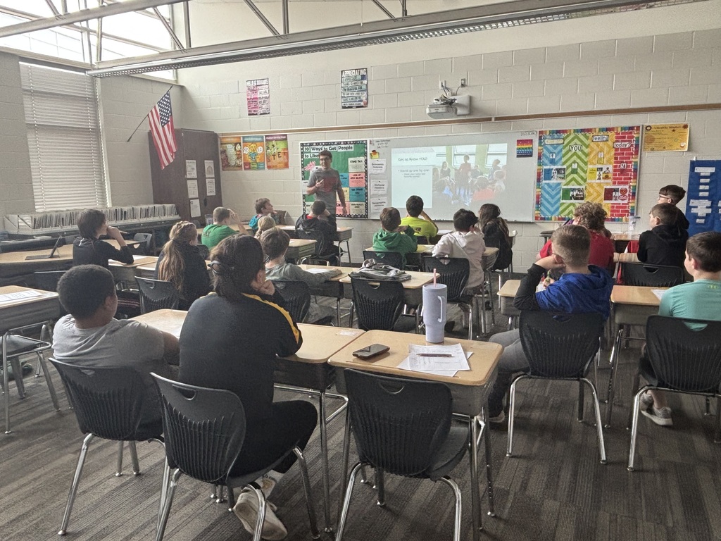 Middle school students seated at desks watching a teacher present at the front of the classroom using a projected slide.