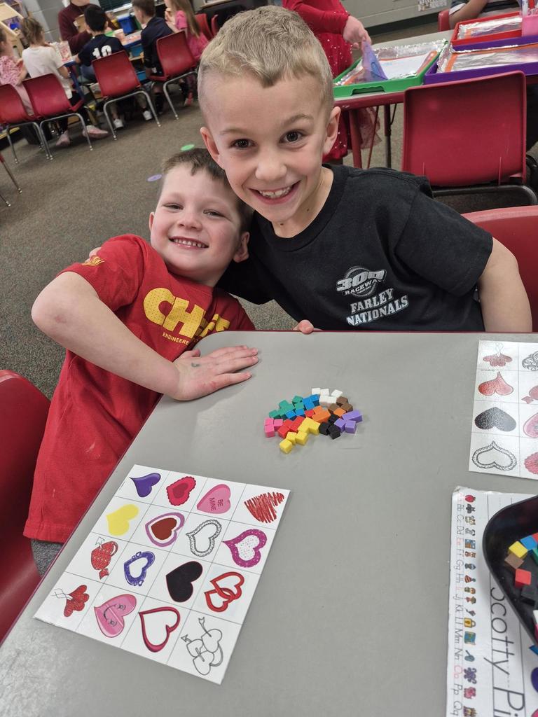 Two young boys smiling at a table with heart-themed bingo cards and colorful counting cubes.